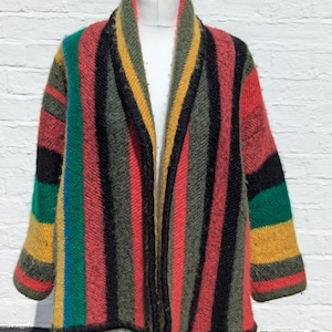 Op de afbeelding: Een kleurrijk gestreept vest in rood, groen, geel en zwart. De open trui wordt getoond op een witte paspop tegen een witte bakstenen muur. De dikke knitwear heeft een sjaalkraag en lange mouwen, ideaal voor de herfst.