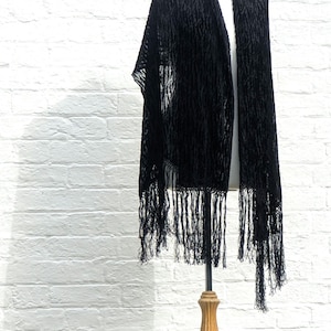 May include: A black shawl featuring a textured, raised pattern throughout the fabric. The rectangular garment is draped over a display form and is finished with long, thin, matching black fringe along the bottom edges, creating a layered look.