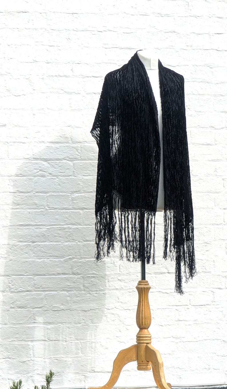 May include: A black shawl featuring a textured, raised pattern throughout the fabric. The rectangular garment is draped over a display form and is finished with long, thin, matching black fringe along the bottom edges, creating a layered look.