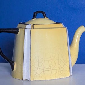 Può includere: Una teiera gialla vintage con manico e coperchio neri. La teiera ha una forma quadrata con dettagli bianchi e neri. La superficie presenta un effetto craquelé, che aggiunge al suo aspetto antico. Il beccuccio è curvo e appuntito.