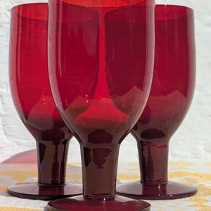 May include: Three matching ruby red glass goblets with smooth, rounded bowls and thick, sturdy stems. Each glass features a wide, flat circular base and a deep, translucent crimson hue that catches the light, creating a uniform set of drinkware.
