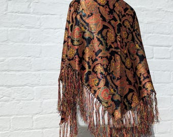 May include: A black shawl featuring a multi-colored paisley pattern in shades of red, gold, and green. The fabric has a subtle sheen and is finished with long, thin, multi-colored fringe along the edges, creating a textured and layered appearance.
