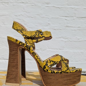 May include: Yellow and black snakeskin-patterned platform sandals with a thick wooden heel and sole. The sandals have a strap around the ankle and over the toes. The heel measures approximately 5 inches.