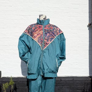 May include: Teal vintage tracksuit with a floral patterned yoke. The jacket has a high collar and a full zip. The trousers are a matching teal colour. The tracksuit is displayed on a mannequin against a white brick wall.