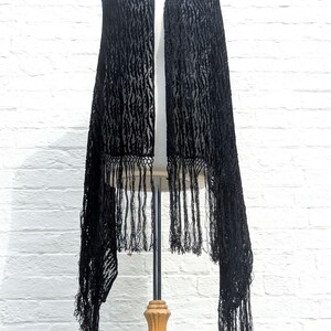 May include: A black velvet shawl featuring a textured, repeating vertical pattern and long, thin fringe along the bottom edge. The fabric has a semi-sheer quality between the raised velvet sections, creating a distinct contrast in light and shadow.