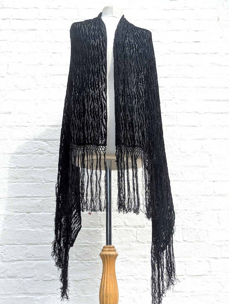 May include: A black velvet shawl featuring a textured, repeating vertical pattern and long, thin fringe along the bottom edge. The fabric has a semi-sheer quality between the raised velvet sections, creating a distinct contrast in light and shadow.