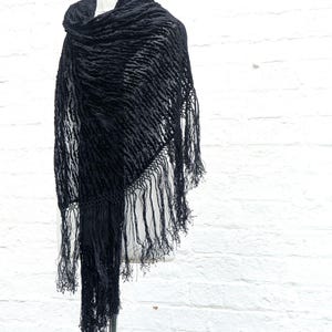 May include: A black triangular shawl featuring a textured, raised velvet-like pattern across the fabric. The edges of the shawl are finished with long, thin, matching black fringe tassels that hang down from the bottom and sides of the garment.