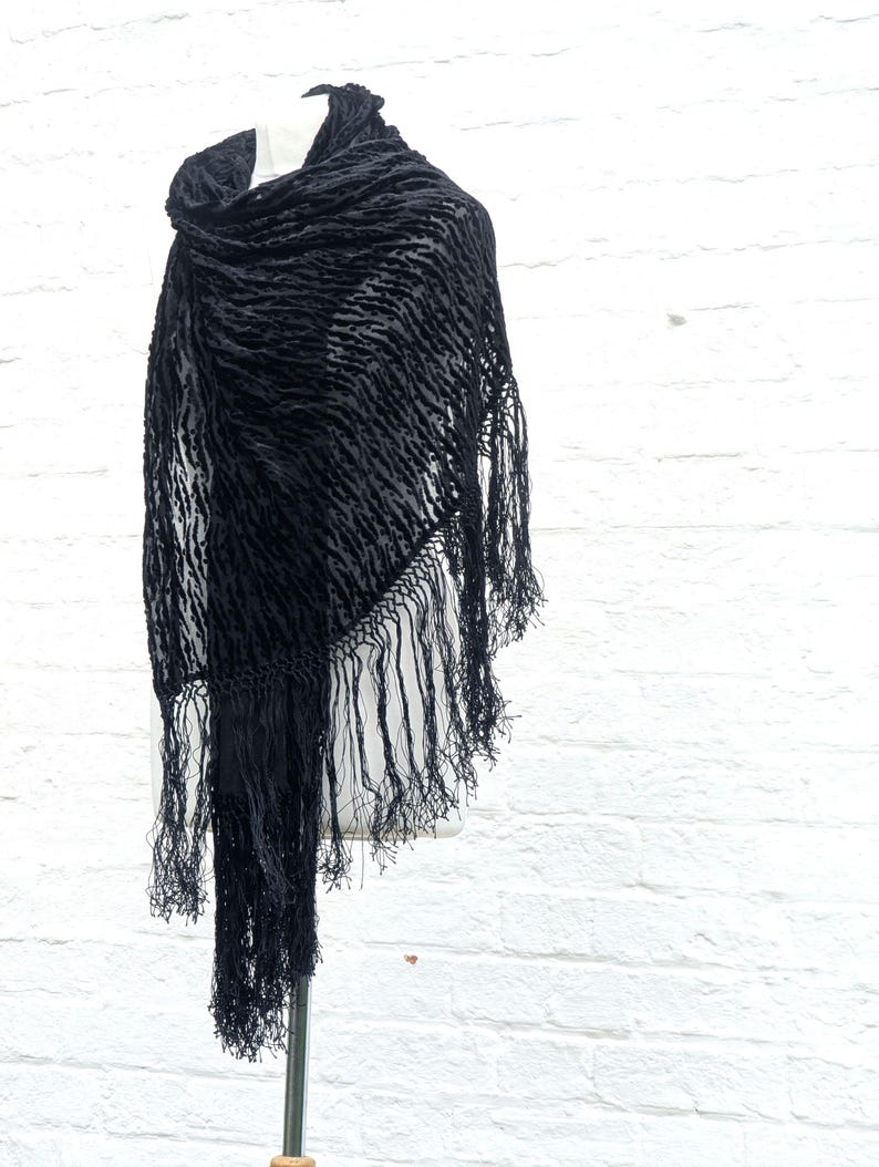 May include: A black triangular shawl featuring a textured, raised velvet-like pattern across the fabric. The edges of the shawl are finished with long, thin, matching black fringe tassels that hang down from the bottom and sides of the garment.