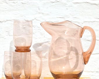 May include: A set of translucent pink glassware consisting of one pitcher with a curved handle and spout, and three matching tumblers. Each piece features a smooth, rounded base and a consistent light pink tint throughout the glass material.