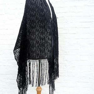 May include: A black shawl featuring a textured, semi-sheer burnout pattern with a repeating vertical design. The garment is finished with long, thin black fringe along the bottom edge, creating a draped silhouette that hangs from a display form.