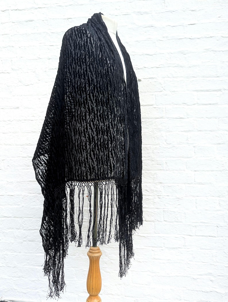 May include: A black shawl featuring a textured, semi-sheer burnout pattern with a repeating vertical design. The garment is finished with long, thin black fringe along the bottom edge, creating a draped silhouette that hangs from a display form.