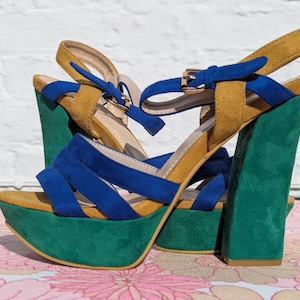 May include: Platform sandals in a vibrant color palette. The shoes feature a green platform sole and heel, with mustard yellow and blue suede straps. The ankle strap has a gold buckle. These bold sandals are a statement piece.