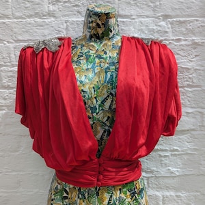 May include: A bright red top with a draped design and short sleeves. The top has a cinched waist with three buttons and is displayed on a mannequin with a patterned fabric. The shoulders are embellished with silver sequins.