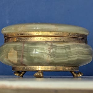 May include: A small, oval-shaped onyx box with a lid. The box is primarily green with gold-colored metal accents around the lid, base, and feet. The box rests on three small, claw-shaped feet.