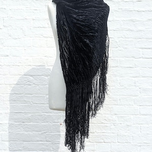 May include: A black triangular shawl featuring a textured, crinkled velvet fabric with a subtle pattern. The garment is draped over a mannequin and is finished with long, thin, matching black fringe along the edges, creating a layered appearance.