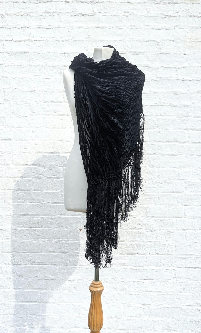 May include: A black triangular shawl featuring a textured, crinkled velvet fabric with a subtle pattern. The garment is draped over a mannequin and is finished with long, thin, matching black fringe along the edges, creating a layered appearance.