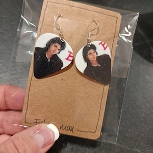 Könnte beinhalten: Ein Paar Ohrringe mit einem Porträt von Michael Jackson. Die Ohrringe sind wie Plektren geformt und haben einen weißen Hintergrund und goldfarbene Haken. Der Buchstabe "E" ist rot auf den Ohrringen aufgedruckt.