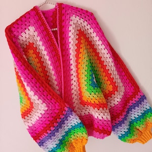 May include: A colourful, handmade crochet cardigan in rainbow hues, including pink, red, orange, yellow, green, and blue. The open-front sweater features a granny square design with long, puffy sleeves and a bright, cheerful aesthetic.