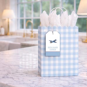 A blue and white checkered gift bag with a tag that reads "Benjamin Cohen" sits on a marble countertop in a kitchen.
