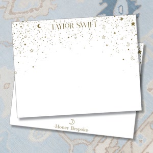 May include: White stationery card with gold stars and crescent moons scattered across the top. The text "TAYLOR SWIFT" is printed in gold at the top of the card. The bottom of the card has the text "Honey Bespoke" printed in gold.