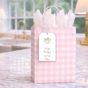 A pink and white checkered gift bag with a birthday tag attached, placed on a marble countertop.