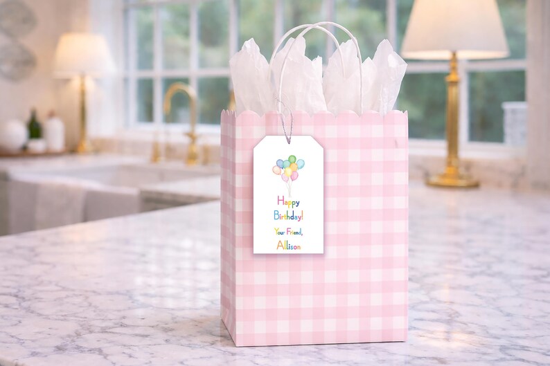 A pink and white checkered gift bag with a birthday tag attached, placed on a marble countertop.