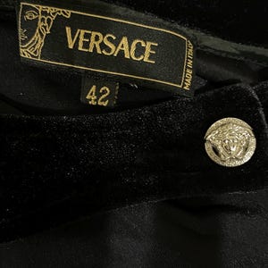 May include: Black velvet trousers with a gold Versace label and a gold button featuring the Versace logo. The label reads "VERSACE" and "MADE IN ITALY". The size tag shows "42".