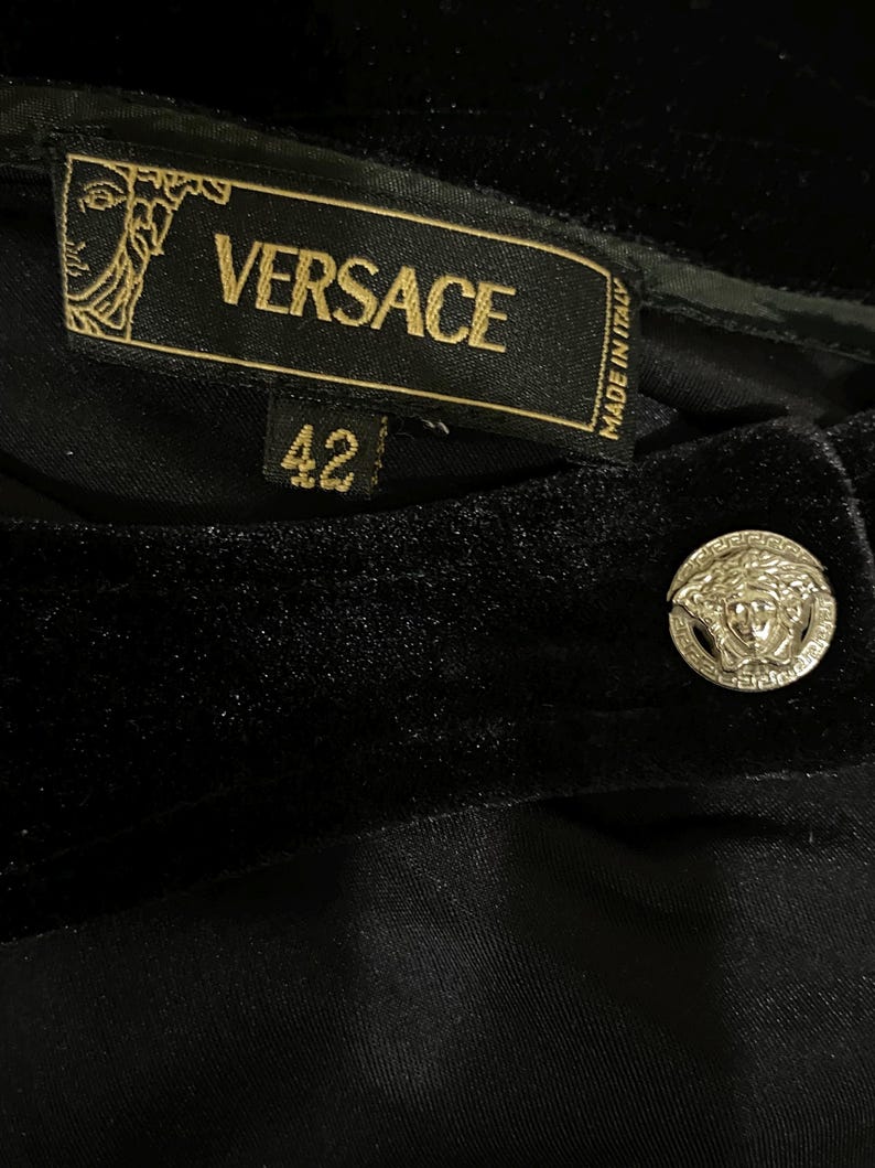 May include: Black velvet trousers with a gold Versace label and a gold button featuring the Versace logo. The label reads "VERSACE" and "MADE IN ITALY". The size tag shows "42".