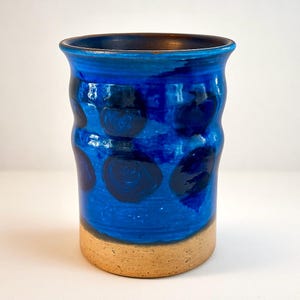 以下が含まれることがあります： 鮮やかな青色の釉薬と濃い青色の水玉模様が特徴の手作りのセラミック花瓶。 花瓶は円筒形で、自然な素焼きのベージュ色のベースが付いています。 内側は濃い青色です。