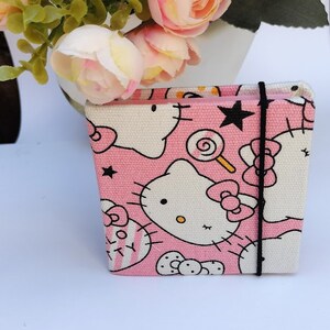 Pode incluir: Um caderno de tela rosa com um design Hello Kitty. O caderno tem um fecho de elástico preto e apresenta um rosto branco Hello Kitty com um laço rosa e uma estrela preta.