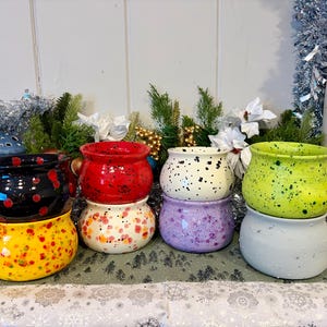 Puede incluir: Seis macetas de cerámica en varios colores, incluyendo negro, rojo, blanco, morado, verde y amarillo. Cada maceta tiene un diseño moteado y está apilada sobre otra maceta. El fondo presenta decoraciones navideñas.