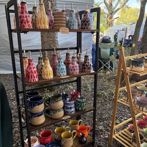 Puede incluir: Un estante de exhibición de metal negro de cuatro niveles que sostiene varias botellas de aceite de cerámica con vertedores en los estantes superiores y tazas y tazones de cerámica en los estantes inferiores. Las tazas tienen esmaltes.