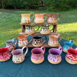 May include: A collection of fourteen ceramic mugs arranged on a tiered wooden stand. Each mug features a speckled base with a distinct colored glaze dripping from the rim, including shades of yellow, orange, red, green, black, pink, purple, and blue.