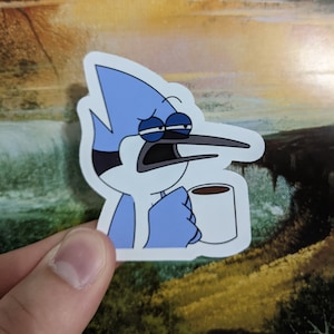 May include: A blue cartoon bird with a grumpy expression is holding a white coffee mug. The bird has a long, pointed beak and large, round eyes. The mug has a brown liquid inside.