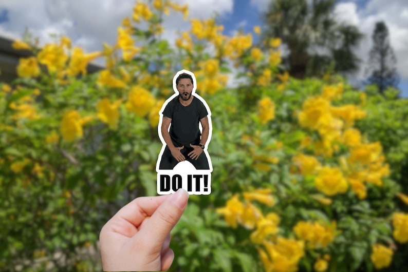 May include: A sticker featuring a man with a beard and a black shirt, with the text "DO IT!" below him. The sticker is held in a hand against a blurred background of yellow flowers.