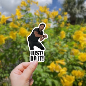 May include: A sticker featuring a man in a black shirt and jeans with the text "JUST! DO IT!" printed below him. The sticker is being held in a hand against a blurred background of yellow flowers.