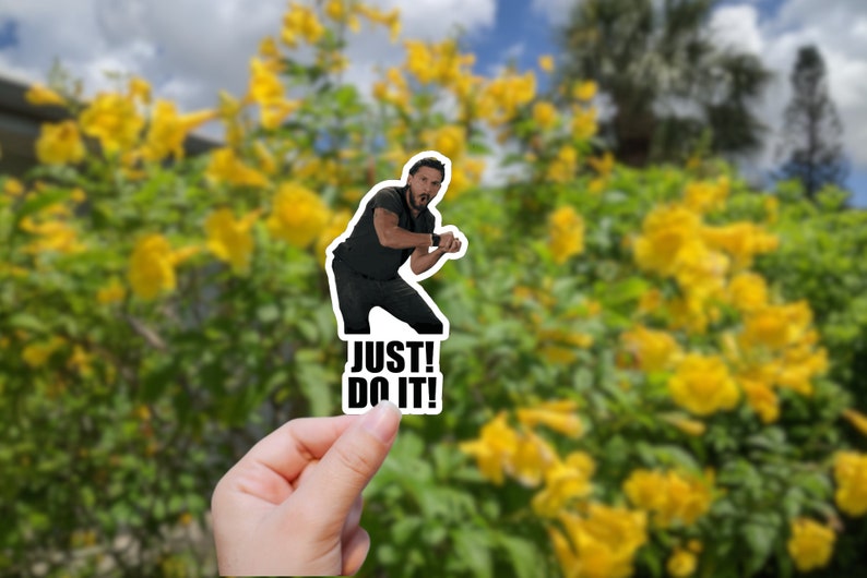 May include: A sticker featuring a man in a black shirt and jeans with the text "JUST! DO IT!" printed below him. The sticker is being held in a hand against a blurred background of yellow flowers.