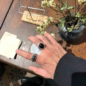 Puede incluir: Se muestran dos anillos en una mano. Uno presenta una piedra negra cuadrada en una banda plateada. El otro tiene un diseño tejido con una piedra negra. Los anillos están en una mano con una manga oscura.