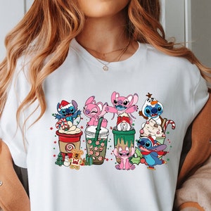 May include: White t-shirt with a colorful graphic of Stitch and Angel characters from the Disney movie Lilo & Stitch. The characters are holding cups of coffee and other beverages, and are decorated with Christmas-themed elements like candy canes and snowflakes.