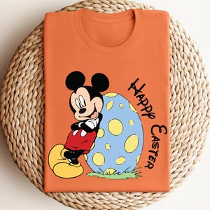 May include: Orange t-shirt with a cartoon image of Mickey Mouse leaning against a blue and yellow Easter egg. The text "Happy Easter" is written vertically on the right side of the shirt.