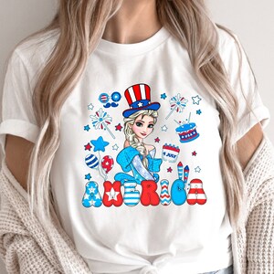 May include: White t-shirt with a cartoon image of a girl wearing a red, white, and blue hat and holding a cup that says "4 JULY". The word "AMERICA" is written in red, white, and blue letters with stars and fireworks around the girl.