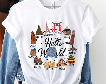 May include: White t-shirt with a graphic design of a globe with various landmarks and the text "Hello World" in a circle around it. The landmarks include the Eiffel Tower, the Tokyo Skytree, the Taj Mahal, the Great Wall of China, and the Statue of Liberty. The text "Hello" is written in multiple languages, including English, French, German, Italian, Spanish, Japanese, and Arabic.