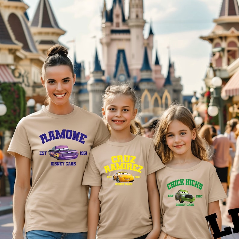 May include: Three people wearing tan t-shirts with Disney Cars graphics. The shirts feature the names "Ramone," "Cruz Ramirez," and "Chick Hicks," along with car illustrations and the text "Disney Cars."