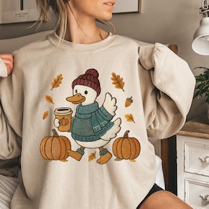 May include: Beige sweatshirt featuring an embroidered duck wearing a knit hat and scarf, holding a coffee cup. The duck is surrounded by pumpkins, leaves, and acorns, creating a fall theme. The sweatshirt has a crew neck and long sleeves.