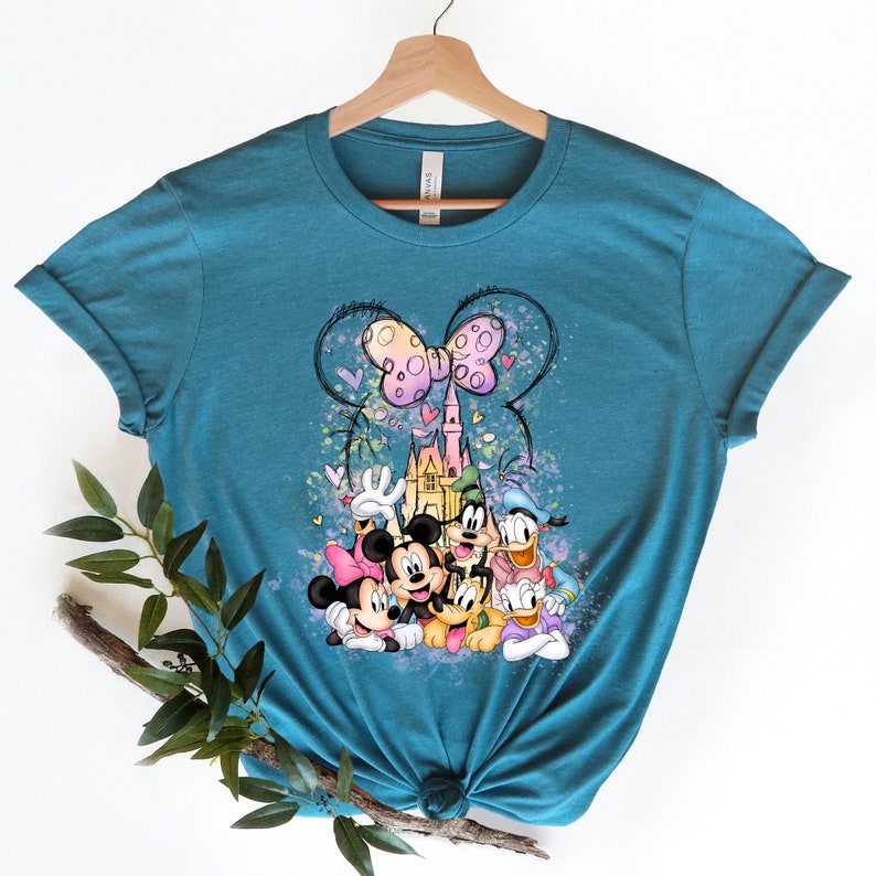May include: Teal blue t-shirt with a colorful graphic print of Mickey Mouse, Minnie Mouse, Donald Duck, Goofy, and Pluto in front of Cinderella's Castle with a pink bow and hearts.