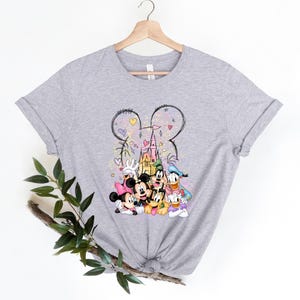 May include: A gray t-shirt with a colorful illustration of Mickey Mouse, Minnie Mouse, Donald Duck, Goofy, and Pluto in front of a pink castle. The illustration is surrounded by hearts and stars.