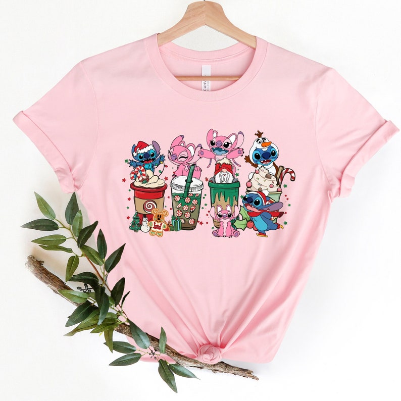 May include: Pink t-shirt with a Christmas themed graphic featuring Stitch and Angel characters from the Disney movie Lilo & Stitch. The characters are holding cups of coffee and surrounded by Christmas decorations.