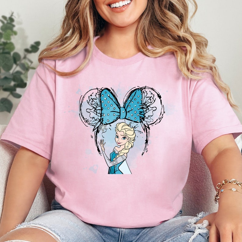 K&ouml;nnte beinhalten: Pinkes T-Shirt mit einer blauen und wei&szlig;en Schleife und einem Cartoon-Bild von Elsa aus Frozen. Die Schleife hat die Form von Mickey-Maus-Ohren.