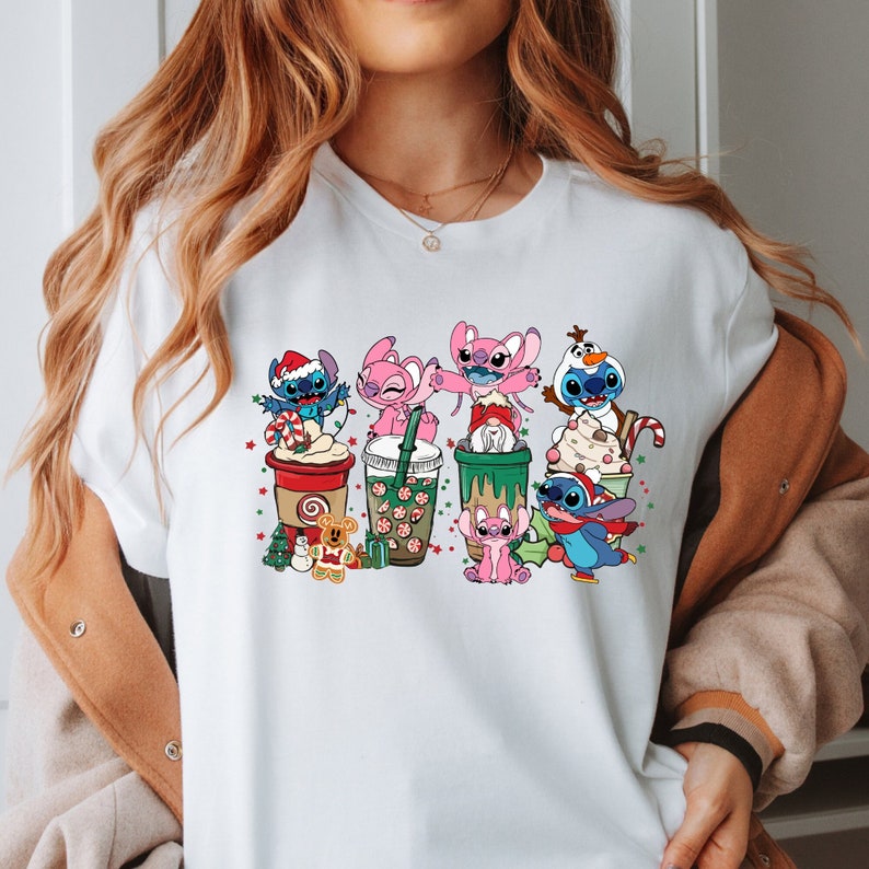 May include: White t-shirt with a colorful graphic of Stitch and Angel characters from the Disney movie Lilo & Stitch. The characters are holding cups of coffee and other beverages, and are decorated with Christmas-themed elements like candy canes and snowflakes.