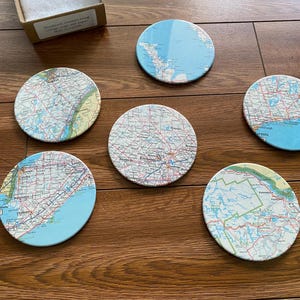 Può includere: Sei sottobicchieri rotondi con un design a mappa. I sottobicchieri sono realizzati in sughero e presentano diverse mappe di varie località. I sottobicchieri sono di colore marrone chiaro con un design a mappa blu e verde.